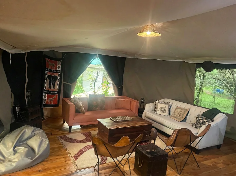 A cozy lounge area inside a safari tent at Kaboso Safari Camp with a rust-colored sofa and a white tufted sofa arranged around an antique wooden chest coffee table playing host to a traditional board game. The room features canvas walls, light wooden floors, an elephant tapestry, and an open tent flap revealing lush green foliage outside.
